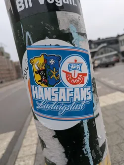 This sticker features the Hansa Fans Ludwigslust emblem. The emblem includes two shields, one with a bull and crown, and the other with a blue design featuring stars and yellow shapes. Next to those is the Hansa Rostock logo with a ship. The text reads 'HANSAFANS Ludwigslust'. The sticker is affixed to a weathered pole with other peeling stickers.