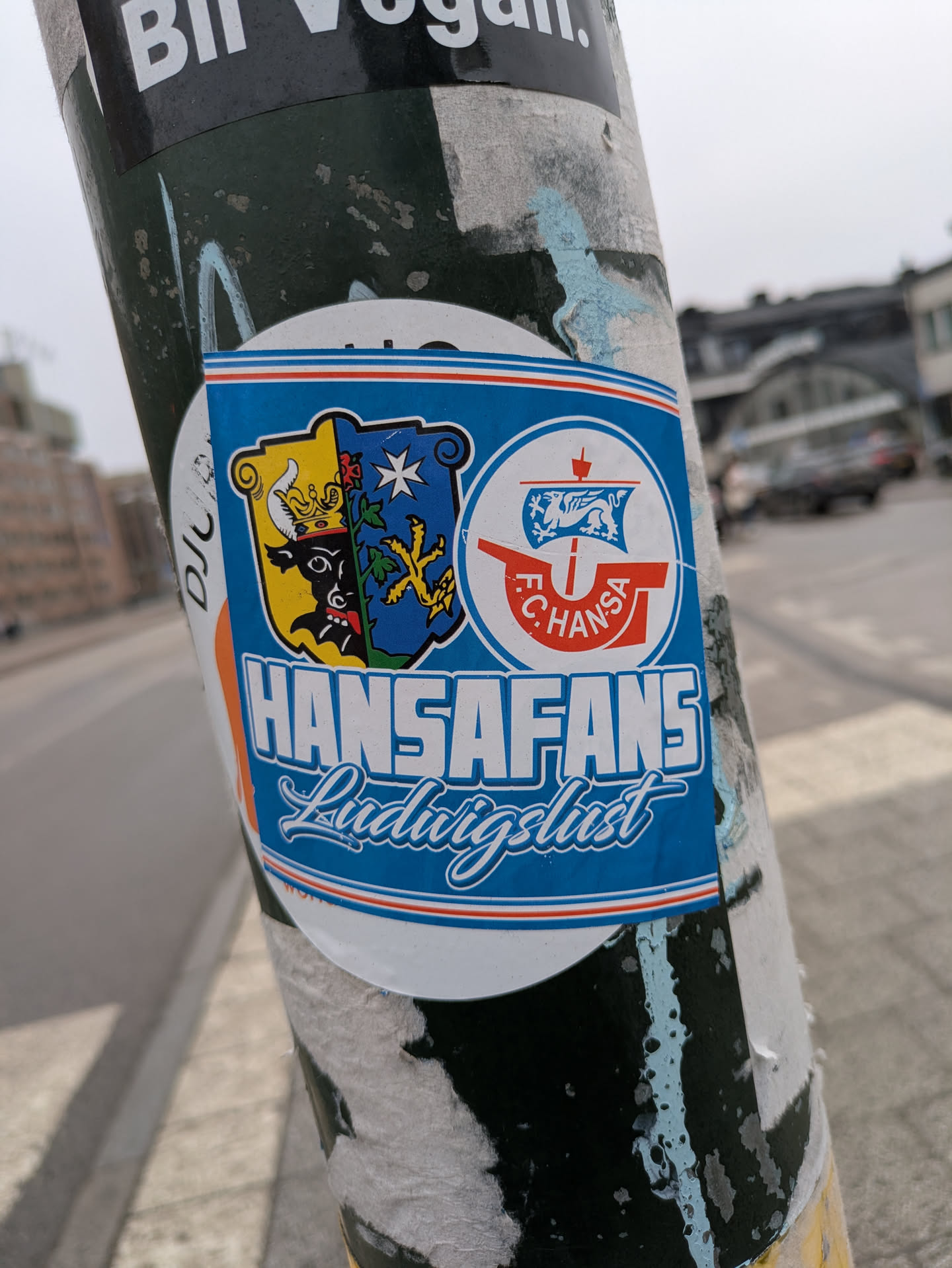 This sticker features the Hansa Fans Ludwigslust emblem. The emblem includes two shields, one with a bull and crown, and the other with a blue design featuring stars and yellow shapes. Next to those is the Hansa Rostock logo with a ship. The text reads 'HANSAFANS Ludwigslust'. The sticker is affixed to a weathered pole with other peeling stickers.