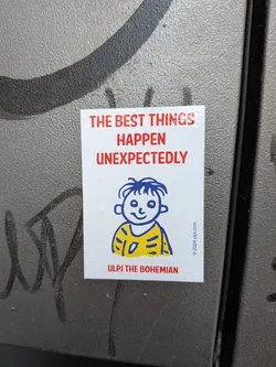 Street sticker A rectangular glossy white sticker applied to a textured, metallic gray surface. At the top, bold red sans-serif text reads 'THE BEST THINGS HAPPEN UNEXPECTEDLY'. Beneath the quote is a simple blue line drawing of a smiling boy character with tousled hair, wearing a yellow shirt marked with blue stripes. Below the character, the name 'ULPI THE BOHEMIAN' is printed in red capital letters. Along the lower right edge, copyright text reads '© 2024 ulpi.com'.