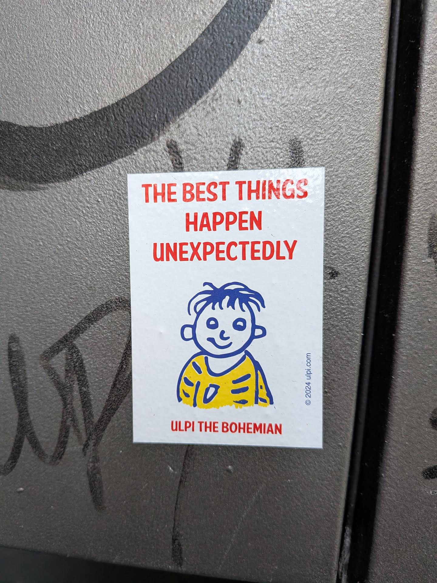 A rectangular glossy white sticker applied to a textured, metallic gray surface. At the top, bold red sans-serif text reads 'THE BEST THINGS HAPPEN UNEXPECTEDLY'. Beneath the quote is a simple blue line drawing of a smiling boy character with tousled hair, wearing a yellow shirt marked with blue stripes. Below the character, the name 'ULPI THE BOHEMIAN' is printed in red capital letters. Along the lower right edge, copyright text reads '© 2024 ulpi.com'.