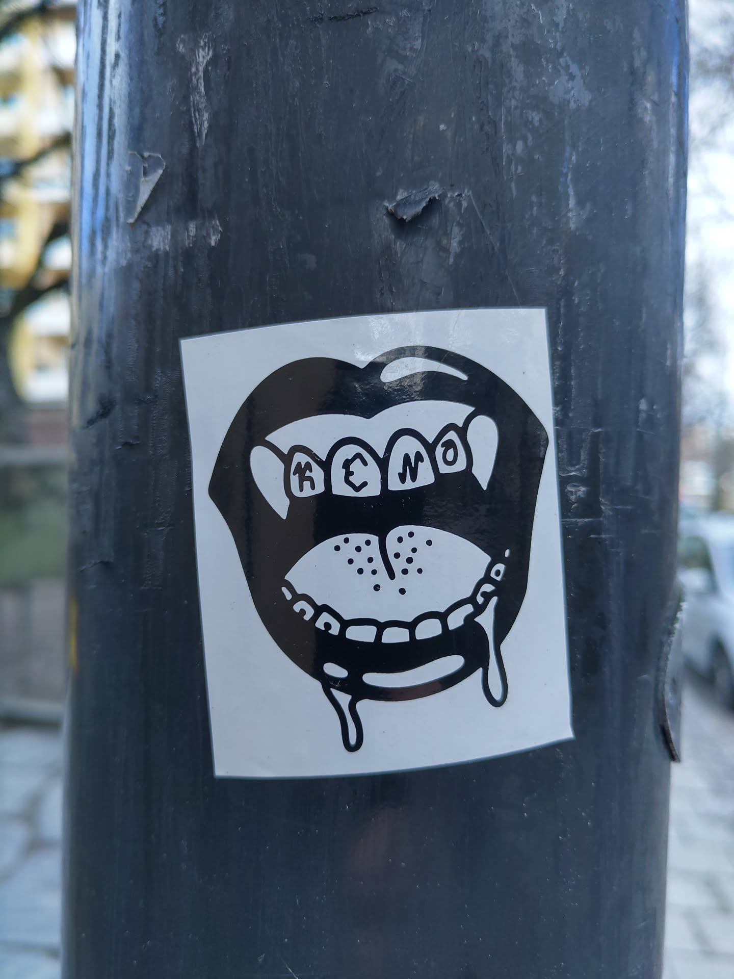 A black and white sticker depicting a cartoonish mouth with teeth dripping. The text 'K3N0' is visible within the mouth.  The sticker is affixed to a dark grey pole.