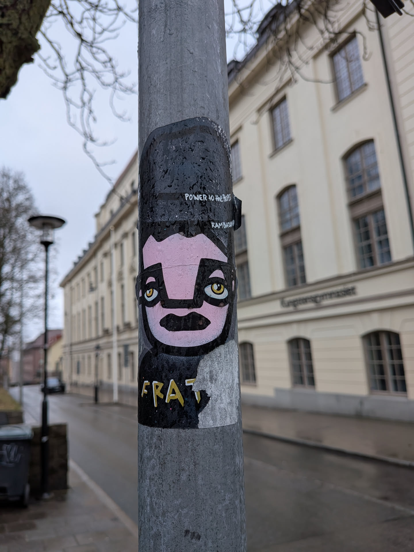 The sticker features a highly stylized, cartoonish character with a prominent pink face, large black-framed eyes with striking yellow irises, and a thick black mustache and mouth. The character wears what appears to be a black cap or has black hair, and a black upper body that transitions into a lighter grey or white lower section. Above the character's head, white text reads 'POWER to the PEOPLE' and 'RAMBAZAM'. Below the character, on the lighter section, the word 'FRAT' is written in yellow letters with a white outline. The overall style is bold and graphic, typical of street art.