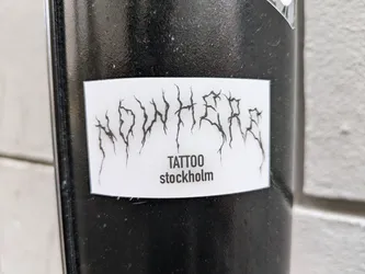 A sticker with the text 'NOWHERE' in a stylized blackletter font, followed by 'TATTOO stockholm' in a smaller font. The sticker is white and rectangular, and is affixed to a black surface.