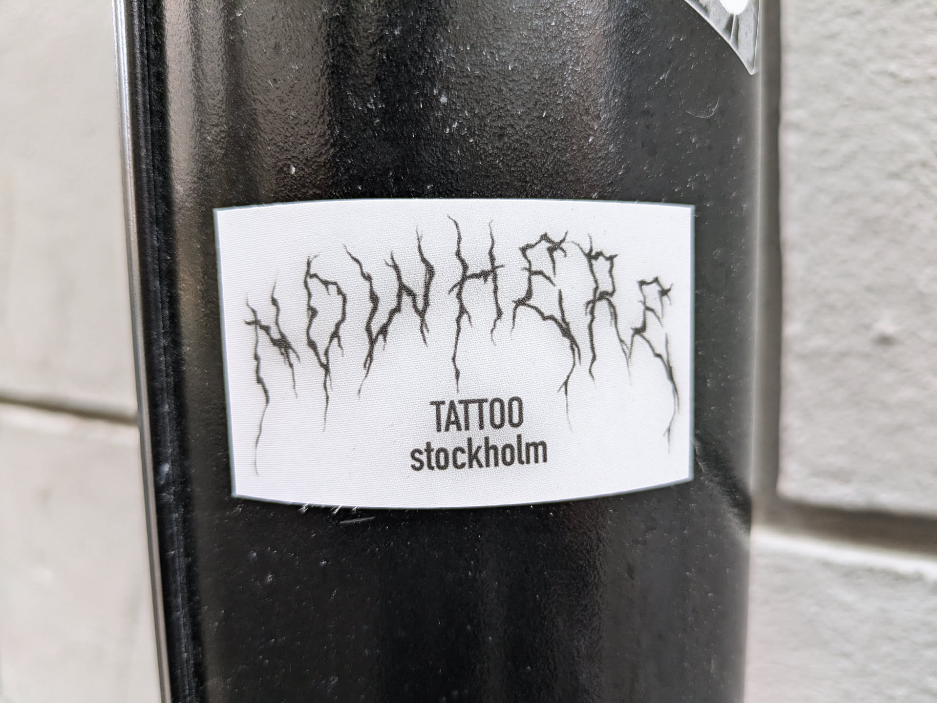 A sticker with the text 'NOWHERE' in a stylized blackletter font, followed by 'TATTOO stockholm' in a smaller font. The sticker is white and rectangular, and is affixed to a black surface.