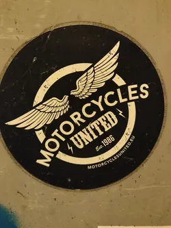 A black and white round sticker with a logo featuring two wings and the text "MOTORCYCLES UNITED" with "Est. 1986" written underneath. The text "MOTORCYCLESUNITED.EU" is written in smaller font at the bottom.
