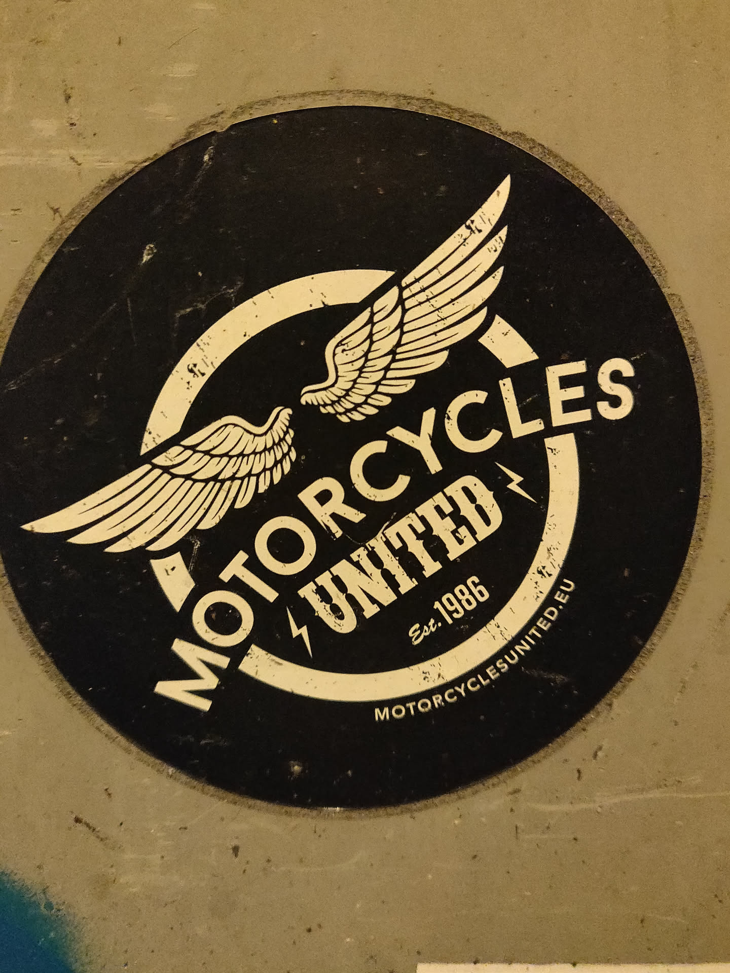 A black and white round sticker with a logo featuring two wings and the text "MOTORCYCLES UNITED" with "Est. 1986" written underneath. The text "MOTORCYCLESUNITED.EU" is written in smaller font at the bottom.