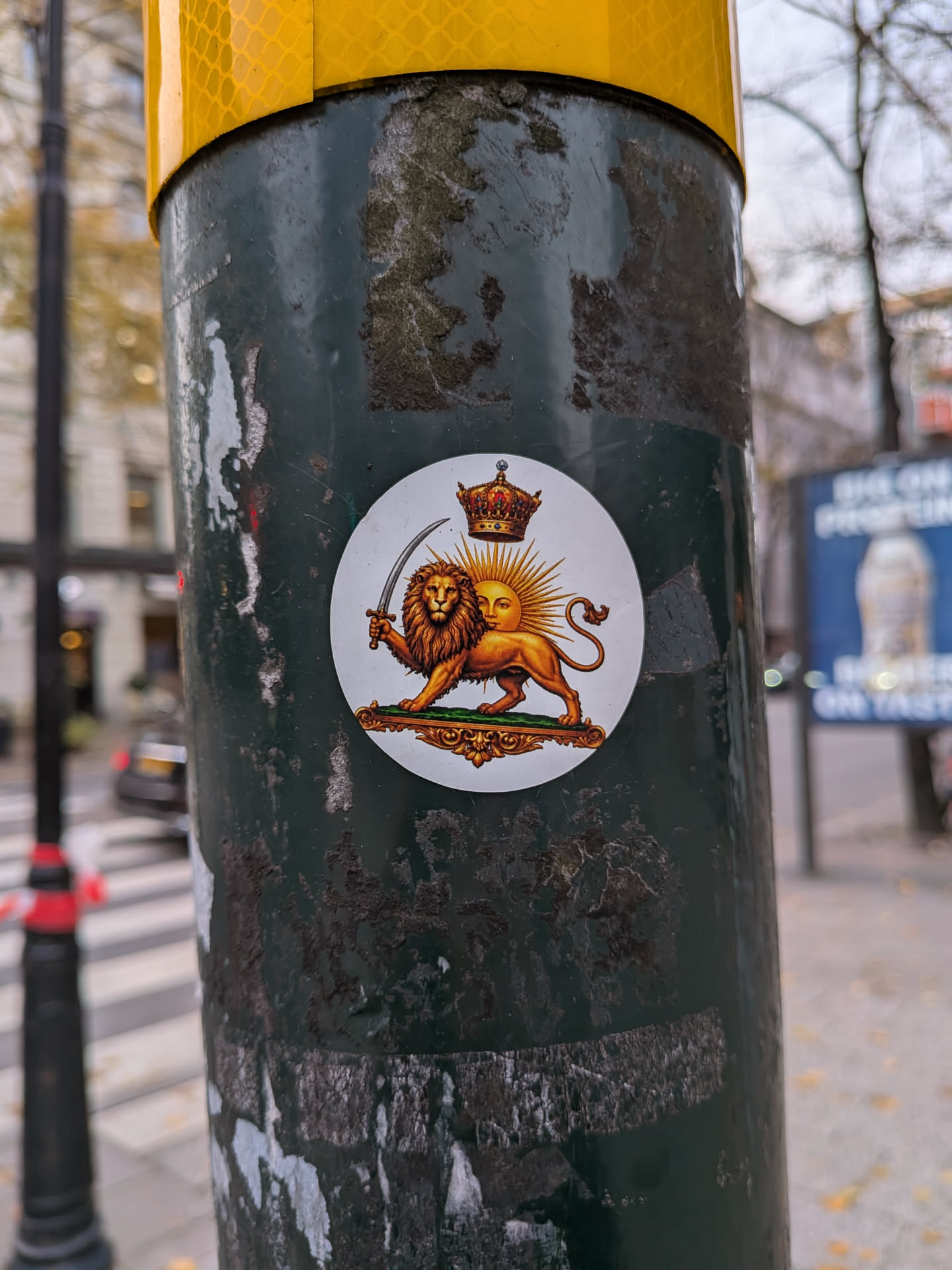 A circular sticker displayed on a dark pole, featuring the classical Iranian Lion and Sun (Shir-o-Khorshid) symbol set against a clean white background. A detailed golden lion stands facing forward while holding a curved shamshir sword in its raised right paw. Behind its back rises a sun with a serene human face radiating golden rays. Floating directly above the sun is an ornate imperial crown detailed with red velvet and precious jewels. The lion rests atop a decorative golden pedestal with a green upper surface.