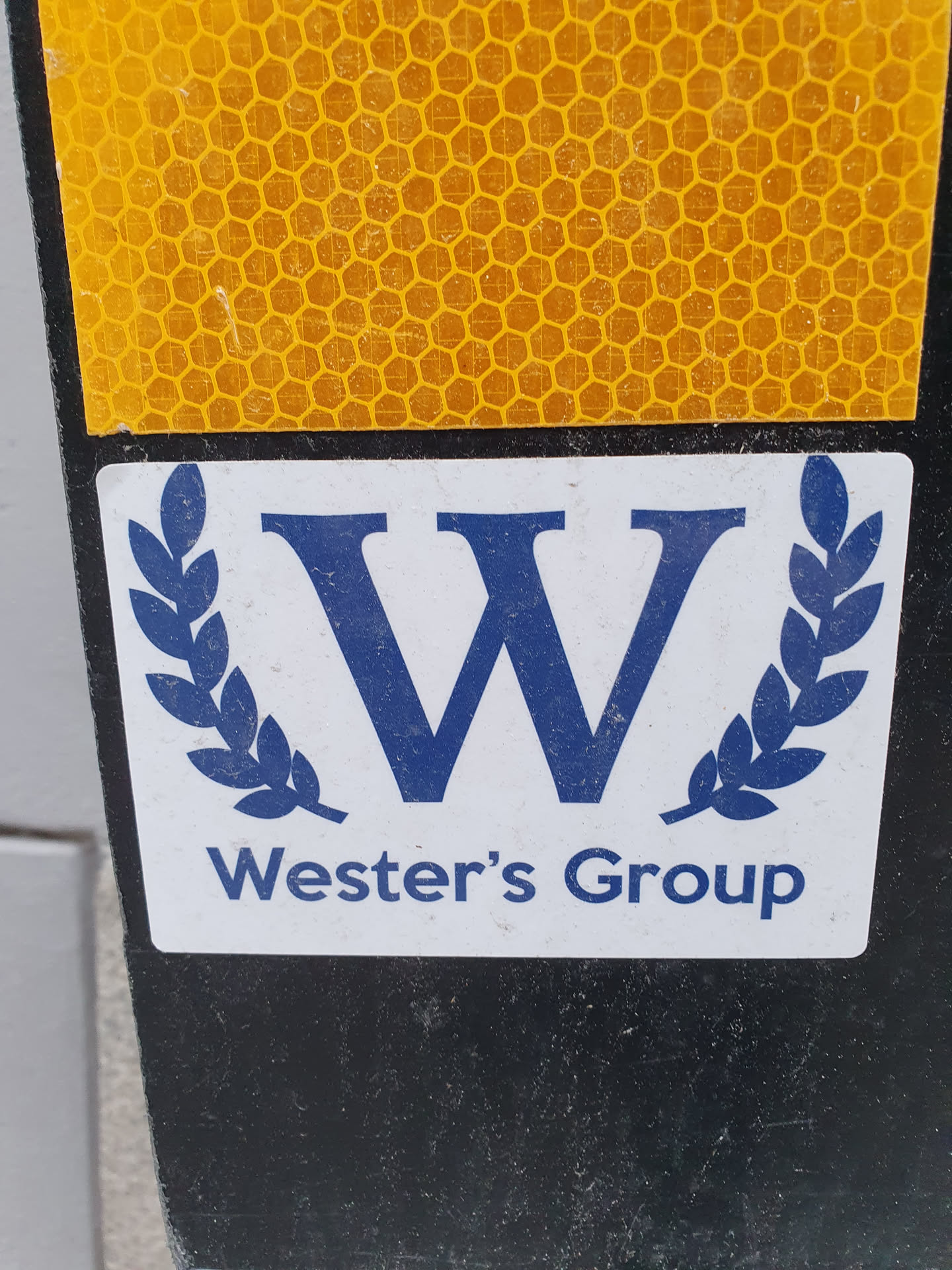 A close-up shot of a Wester's Group logo sticker. The sticker features a blue 'W' surrounded by a laurel wreath, with 'Wester's Group' written underneath. The sticker is affixed to a black surface with a yellow honeycomb pattern visible in the background.