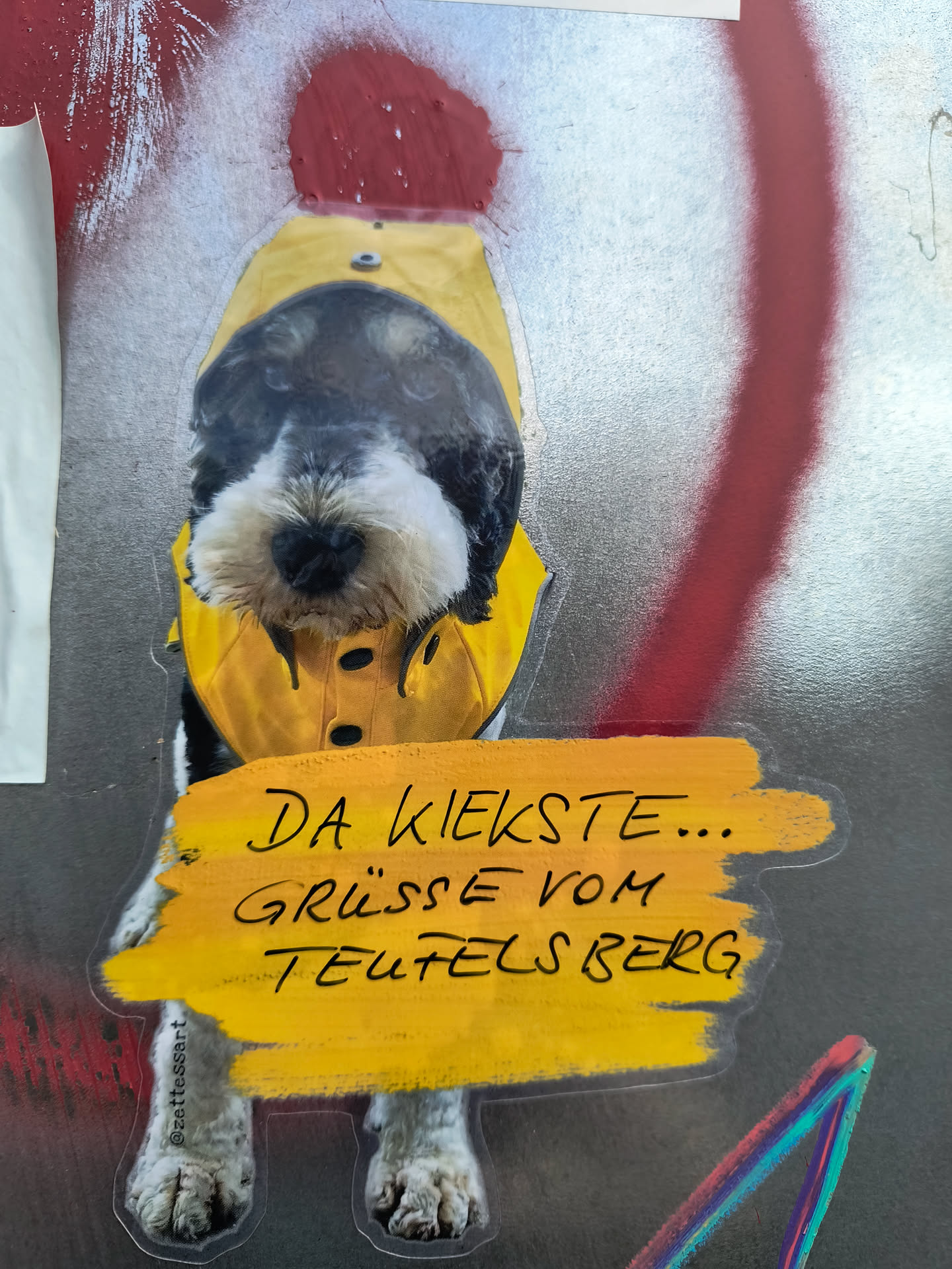 Sticker of a dog wearing a yellow raincoat holding a yellow sign that says: Da kiekste... Grüsse vom Teufelsberg (Here you look... Greetings from Teufelsberg)