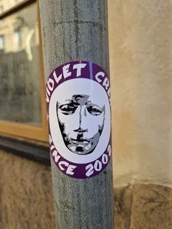 Round sticker with a purple border and the text "Violet Crew Since 2003."  It features a stylized black and white image of a face, possibly a mask, in the center. The sticker is affixed to a metal pole.