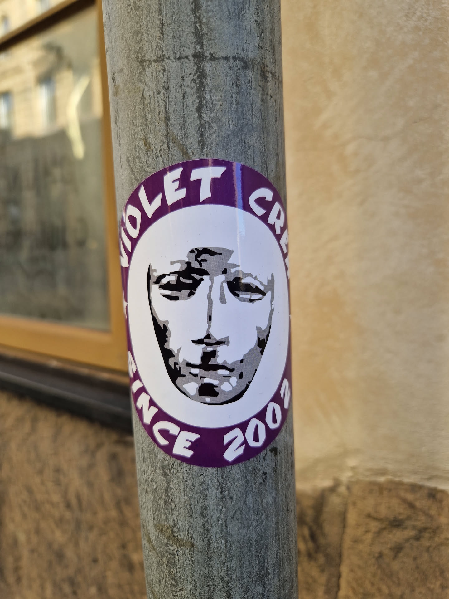 Round sticker with a purple border and the text "Violet Crew Since 2003."  It features a stylized black and white image of a face, possibly a mask, in the center. The sticker is affixed to a metal pole.