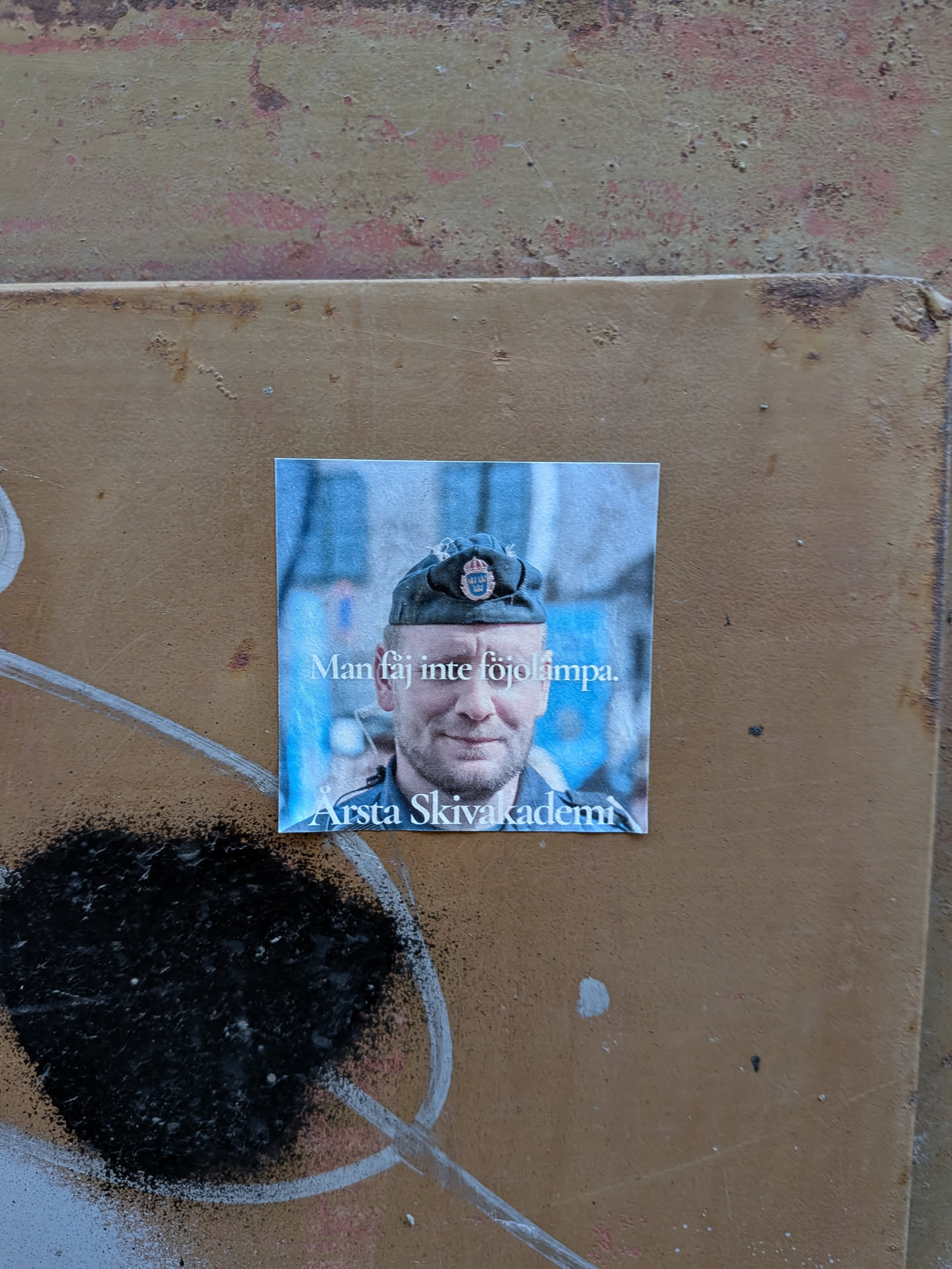 The sticker features a close-up portrait of a smiling man wearing a dark military-style cap with a Swedish emblem. The background is blurred and appears to be an urban setting with blue tones. White text is overlaid on the lower part of the image. The sticker itself is square with slightly rounded corners.