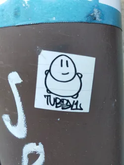 A sticker featuring a simple drawing of a tubby character with a smiling face. The sticker is placed on a brown pole. The artist's tag, TUBBY11, is written underneath the character.
