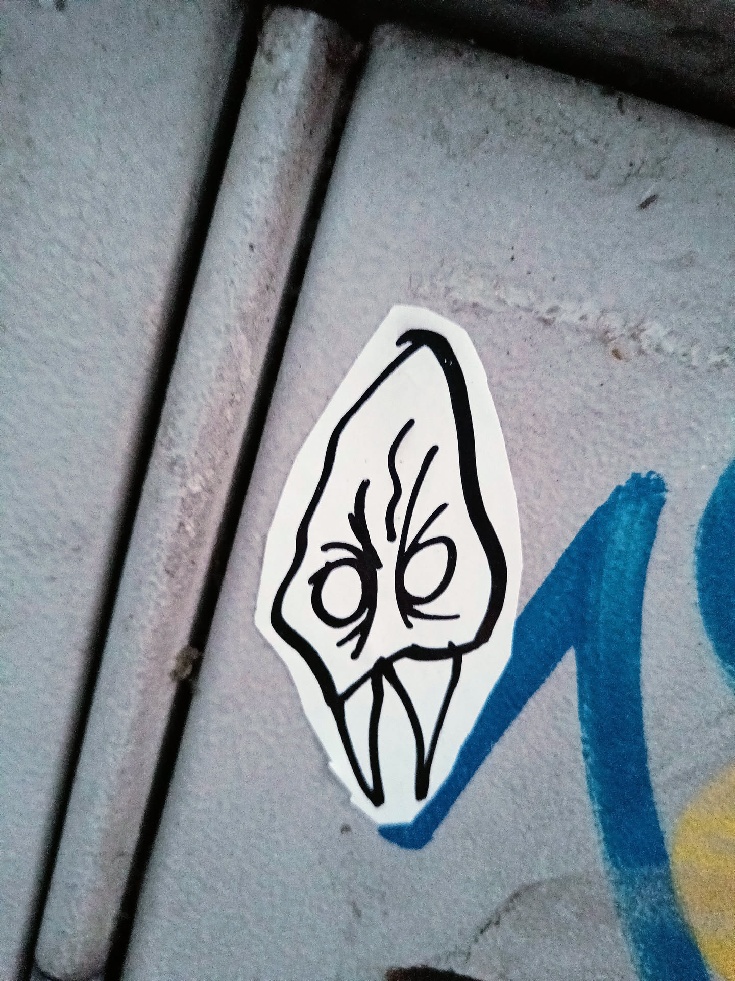 A small, white sticker with a black ink drawing of a creature resembling a bird or a mask. It has large eyes, a beak-like mouth and sharp teeth. The sticker is affixed to a grey surface with some other graffiti.