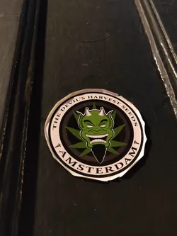 Round sticker featuring a green devil-like face surrounded by marijuana leaves. The text on the sticker says "The Devil's Harvest Seeds Amsterdam".