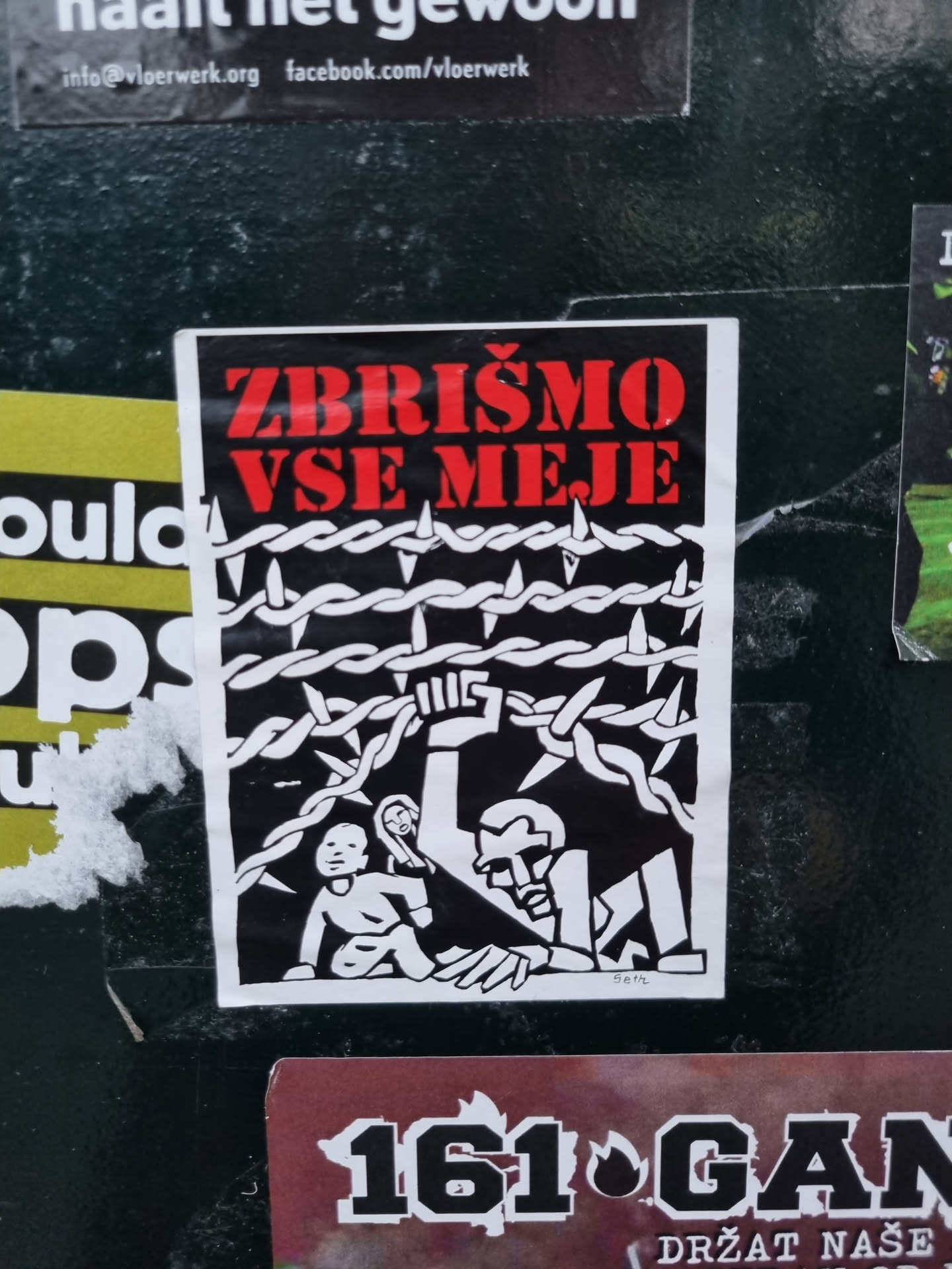 A black and white sticker with red text. The text in the sticker says "Zbrišimo vse meje", which translates to "Let's erase all borders". The image depicts a person reaching over a barbed wire fence to help a child. 