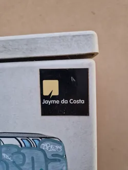 A black sticker with a yellow logo and the name Jayme da Costa printed in white. The sticker is placed on a light beige wall.