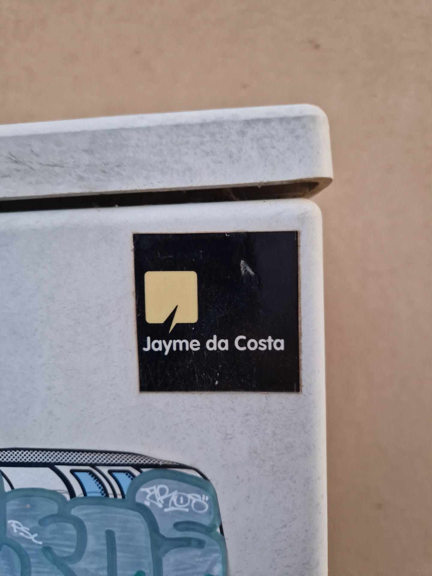 A black sticker with a yellow logo and the name Jayme da Costa printed in white. The sticker is placed on a light beige wall.