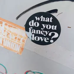 A round black and white sticker with the text "What do you fancy? Love?" in a bold sans-serif font. The sticker is affixed to a light gray surface.