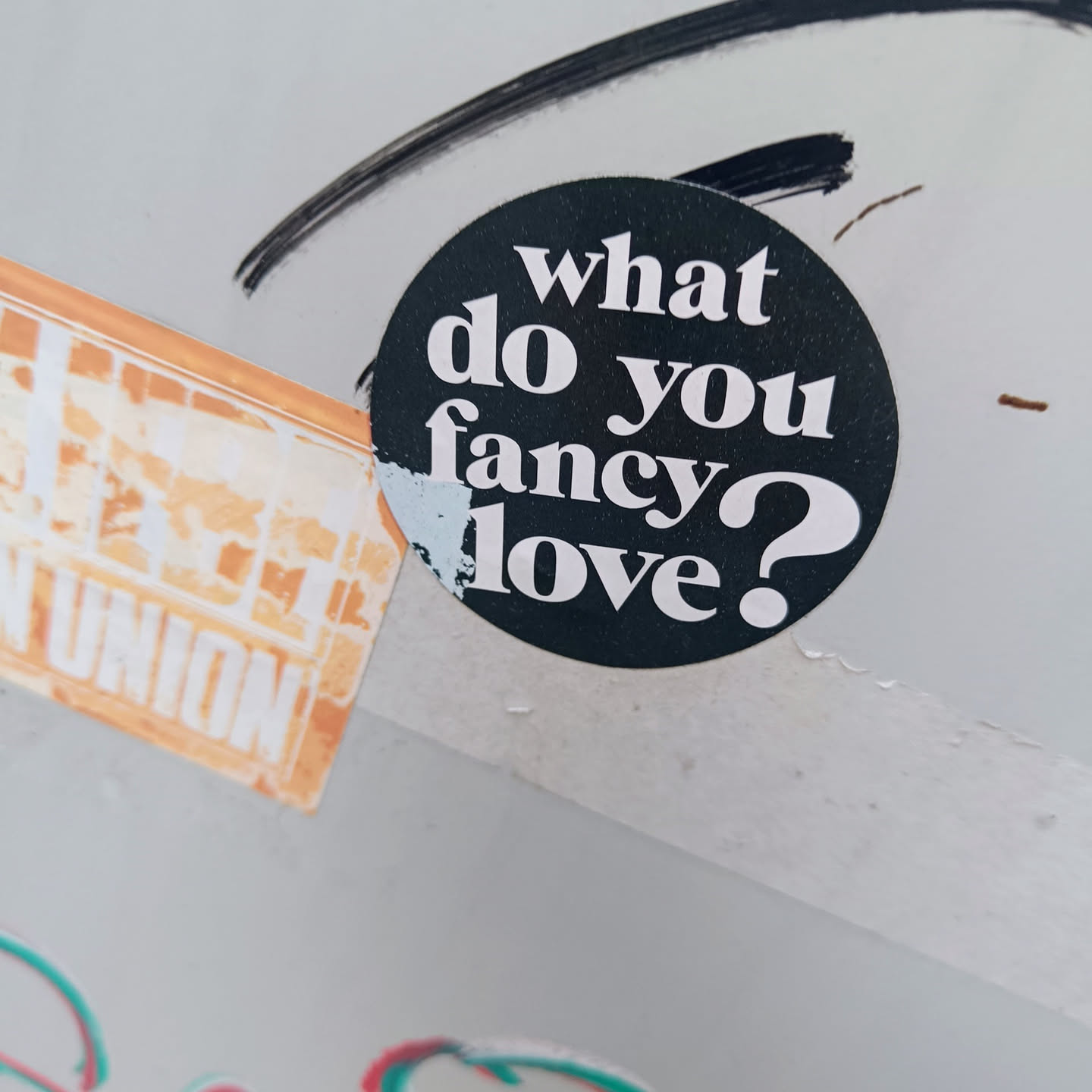 A round black and white sticker with the text "What do you fancy? Love?" in a bold sans-serif font. The sticker is affixed to a light gray surface.