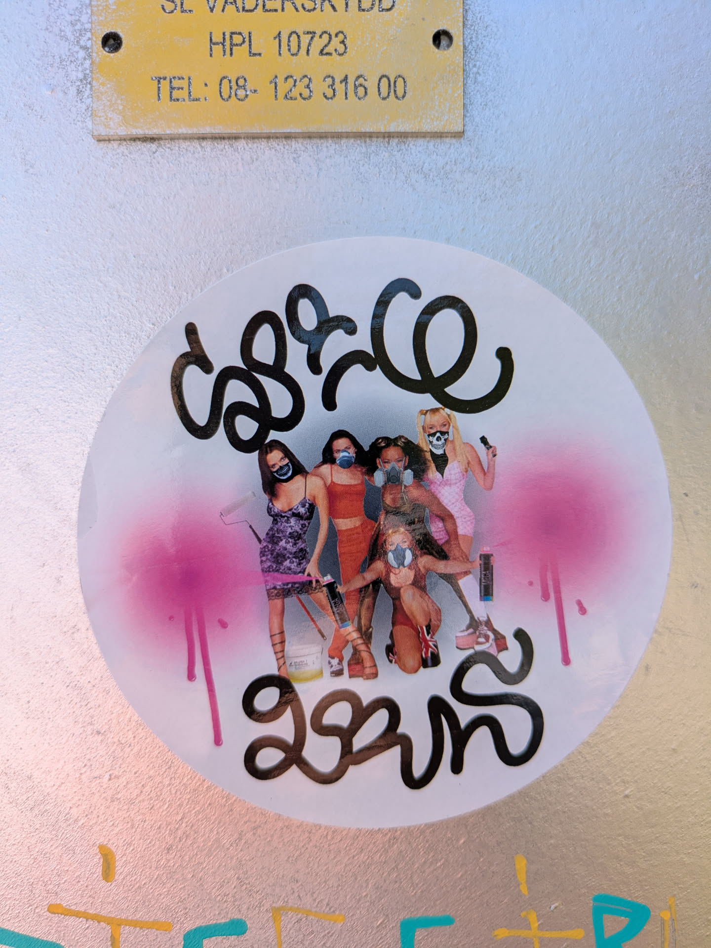 This round sticker is pasted on a metal surface. It features a collage of women wearing masks and safety goggles. Some are holding tools and standing in suggestive poses. Above and below the image, there is graffiti-style text spray-painted in black. There is pink paint splashed on the left side of the sticker.