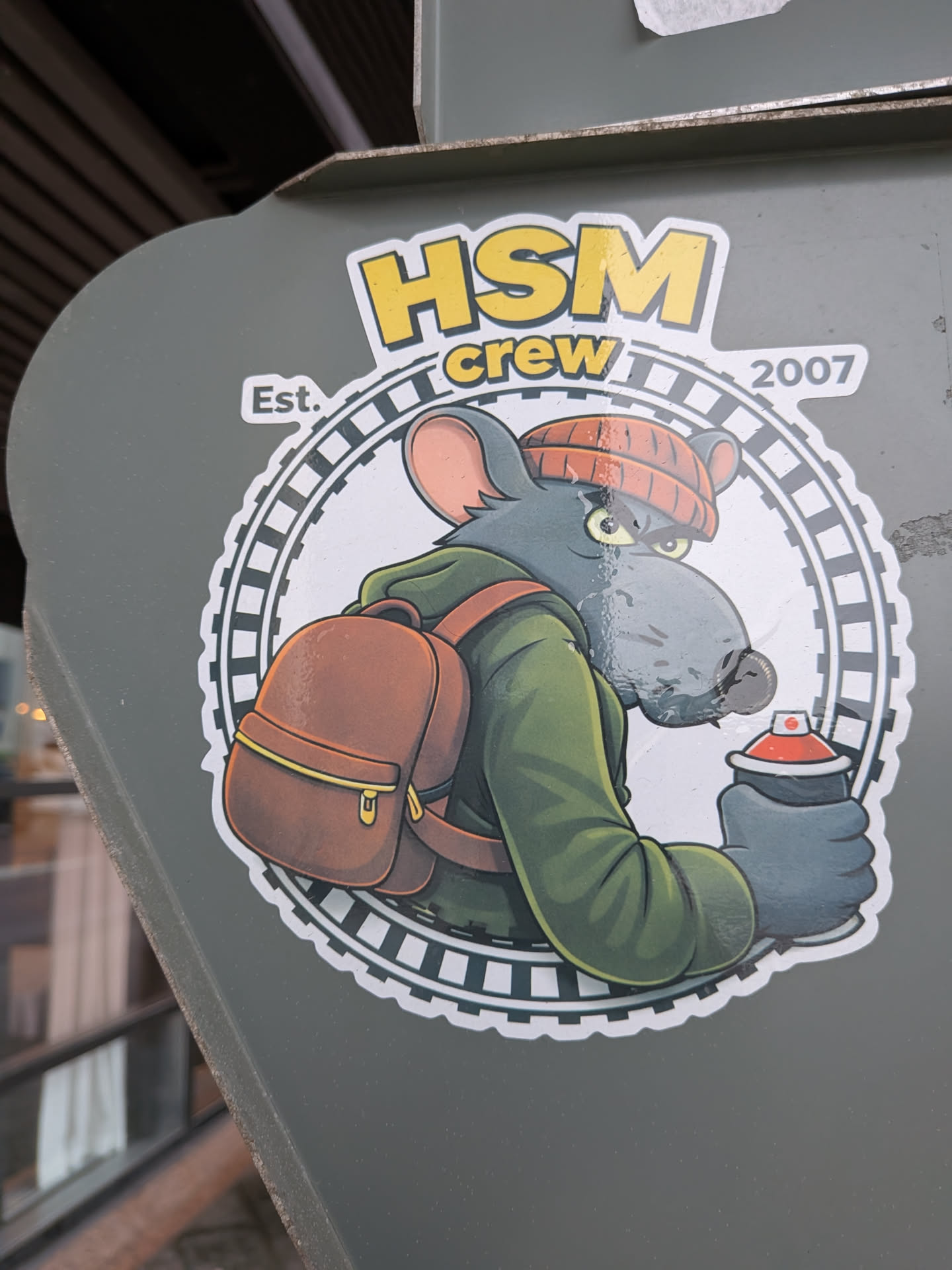 A die-cut sticker featuring a stylized cartoon grey rat glancing back with a focused glare. It wears an orange knit beanie, a green hooded jacket, and a brown backpack, while holding a black spray paint can with a red top. The character is enclosed within a circular emblem surrounded by train track motifs. Bold yellow lettering at the top reads 'HSM crew' flanked by 'Est.' on the left and '2007' on the right.