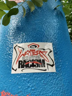 A sticker expressing solidarity against racism. It features stylized graffiti-like lettering.