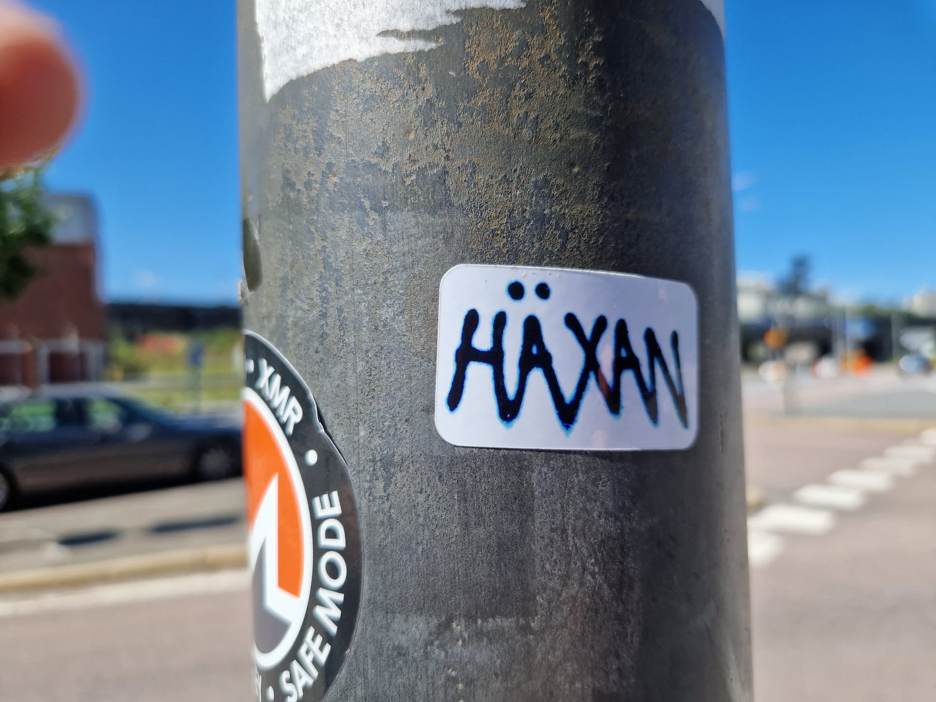 A small white sticker with the word Häxan written in dark blue, slightly stylized lettering. The sticker is affixed to a grey metal pole outdoors.