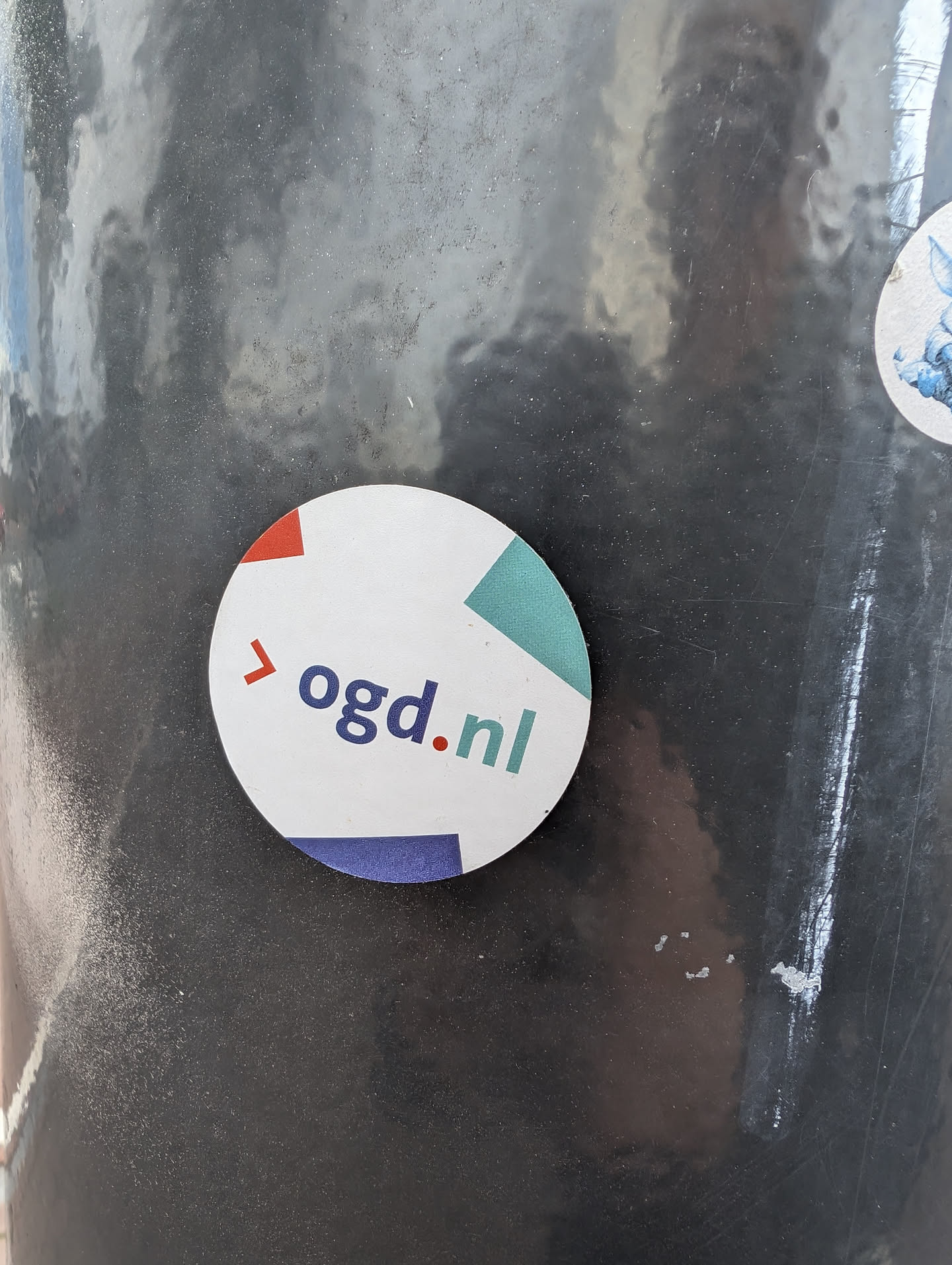 A round sticker with the text 'ogd.nl' and a geometric design in red, teal and blue.  The sticker is affixed to a dark grey surface.
