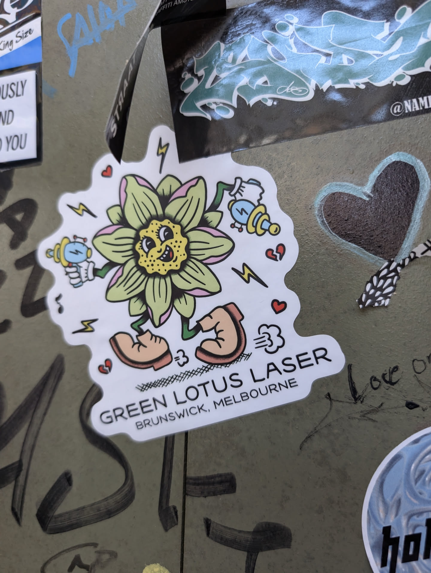 A sticker featuring a whimsical cartoon illustration of a flower character holding ray guns. The text on the sticker reads "GREEN LOTUS LASER" and "BRUNSWICK, MELBOURNE".