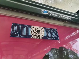 A sticker featuring the numbers 2003 and a skull graphic. The sticker is dark blue and light blue, and is affixed to a pink surface. 