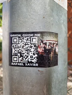 A sticker featuring a QR code, the text "Orion, Guide Me" and "Rafael Xavier", and an image of a group of people. The sticker is affixed to a gray metal pole.