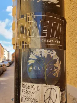 A black and white sticker with the words "TREN CREATIVES" at the top, and a circular sticker with an illustration of white lilies and the text "4 FLIEGEN". There's also a smaller sticker with handwritten text in Swedish and a peace symbol.