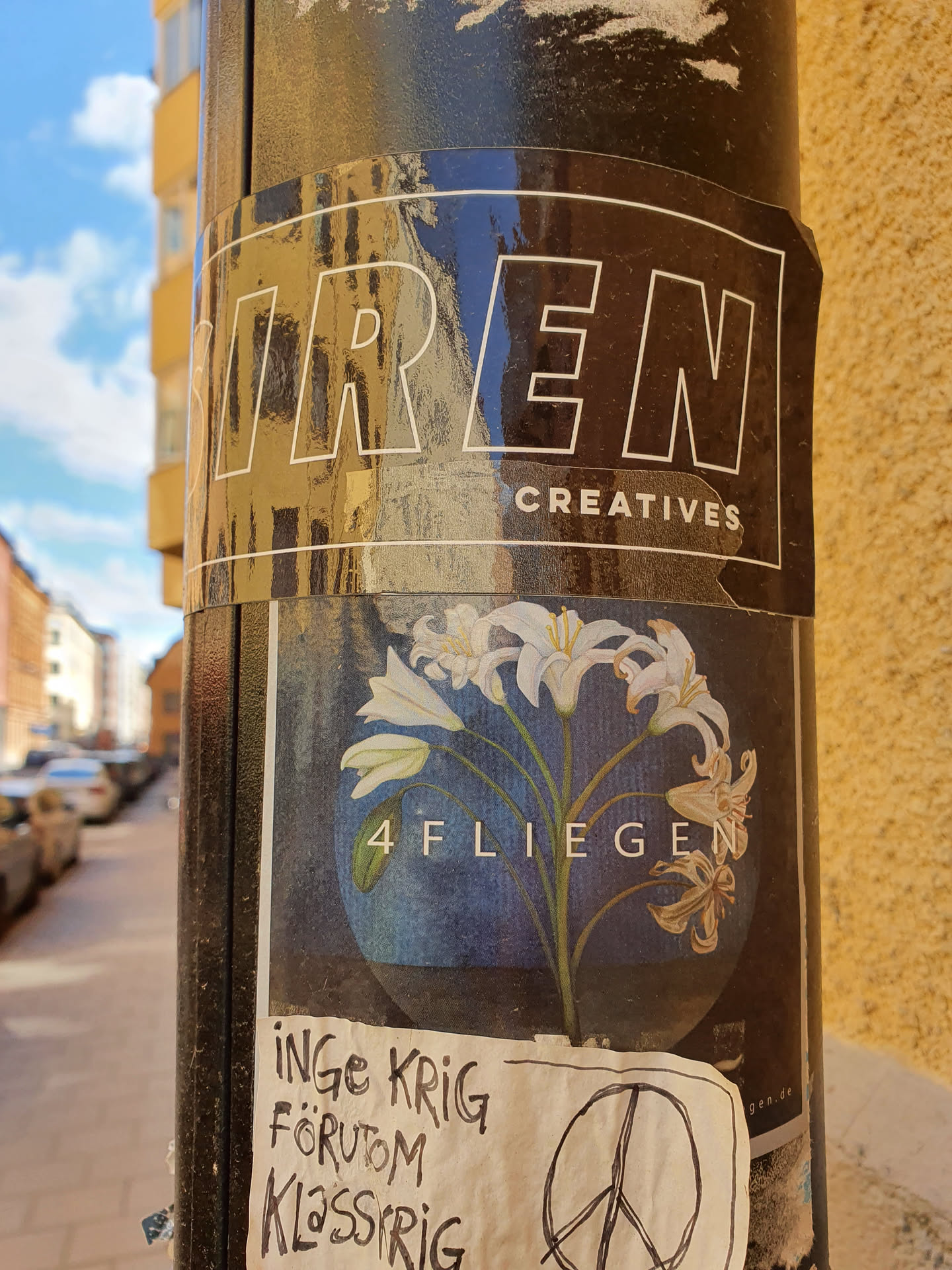 A black and white sticker with the words "TREN CREATIVES" at the top, and a circular sticker with an illustration of white lilies and the text "4 FLIEGEN". There's also a smaller sticker with handwritten text in Swedish and a peace symbol.