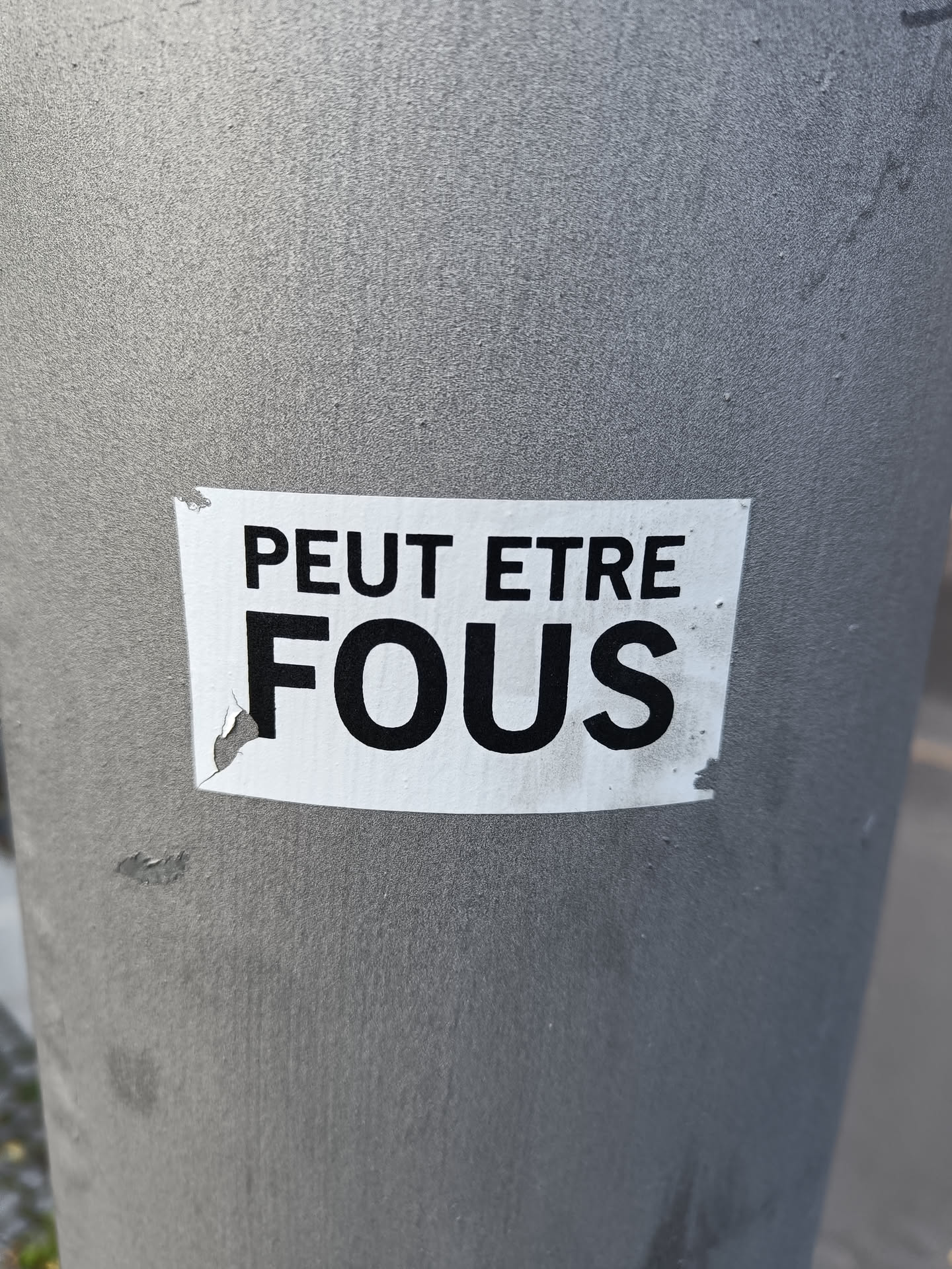 A sticker on a grey pole with the text "Peut être fou(s)" which translates to "Maybe crazy" or "Maybe crazy (plural)" in English. The sticker is slightly damaged.