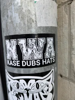 Black and white sticker with the letters NWA in a stylized font, with a paisley-like pattern inside each letter. Below the letters is the text "Kase Dubs Hats".
