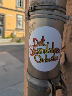 The sticker is white and circular, prominently featuring text in a playful, hand-drawn style. The letters transition from a reddish-orange at the top to a golden-orange at the bottom, with a distinct black outline. The sticker is attached to a greyish metal pipe or drainpipe, which is secured to a building's facade by metal clamps and plastic zip ties. The background shows a blurry street scene with a yellow building.