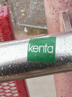 A rectangular sticker with a vibrant green background, featuring the word 'kenta' in clean, white, lowercase sans-serif letters. The sticker appears slightly wrinkled and is covered in numerous small water droplets, indicating it is wet. It is affixed to a reflective, chrome-like metal pole, also covered in water droplets.