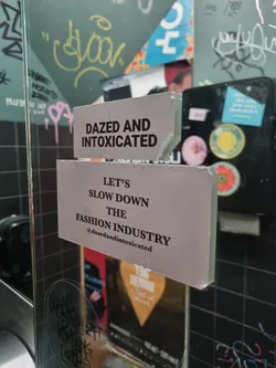 A sticker with a message advocating for a slower fashion industry.  It's been affixed to a glass surface among other stickers and graffiti.
