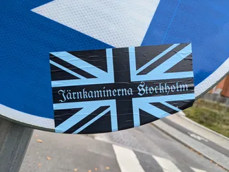 A sticker featuring a Union Jack flag with the text "Järnkaminerna Stockholm" in light blue lettering. The sticker is applied to a blue traffic sign.