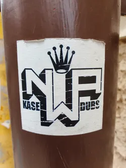 A worn sticker featuring the logo "NP" with "KASE" on the left and "DUBS" on the right, and a crown above it. The sticker is affixed to a brown pole.