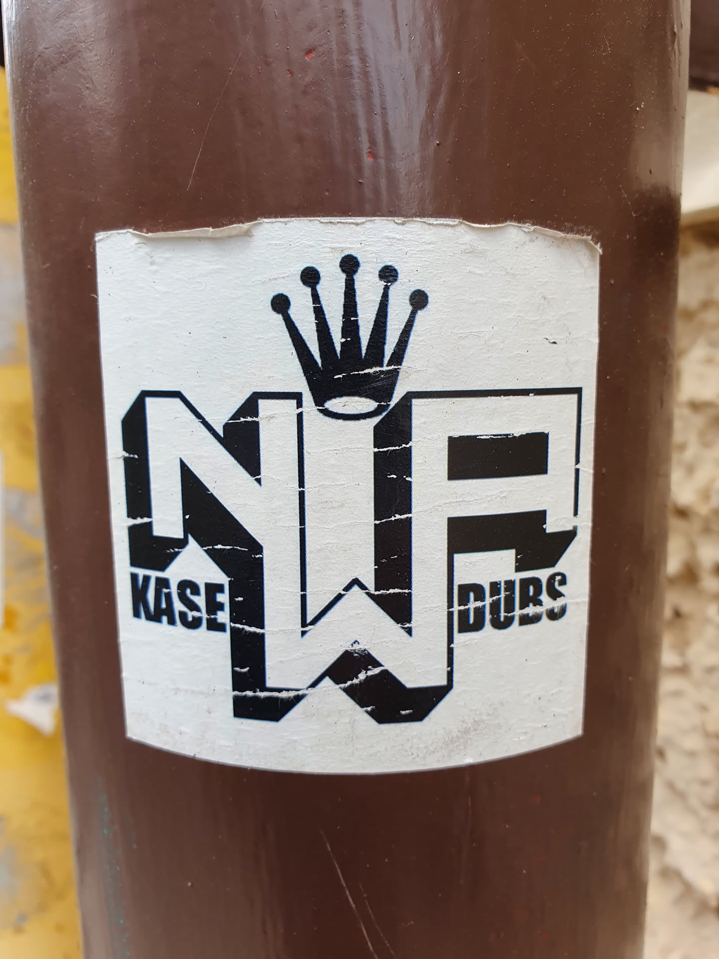 A worn sticker featuring the logo "NP" with "KASE" on the left and "DUBS" on the right, and a crown above it. The sticker is affixed to a brown pole.