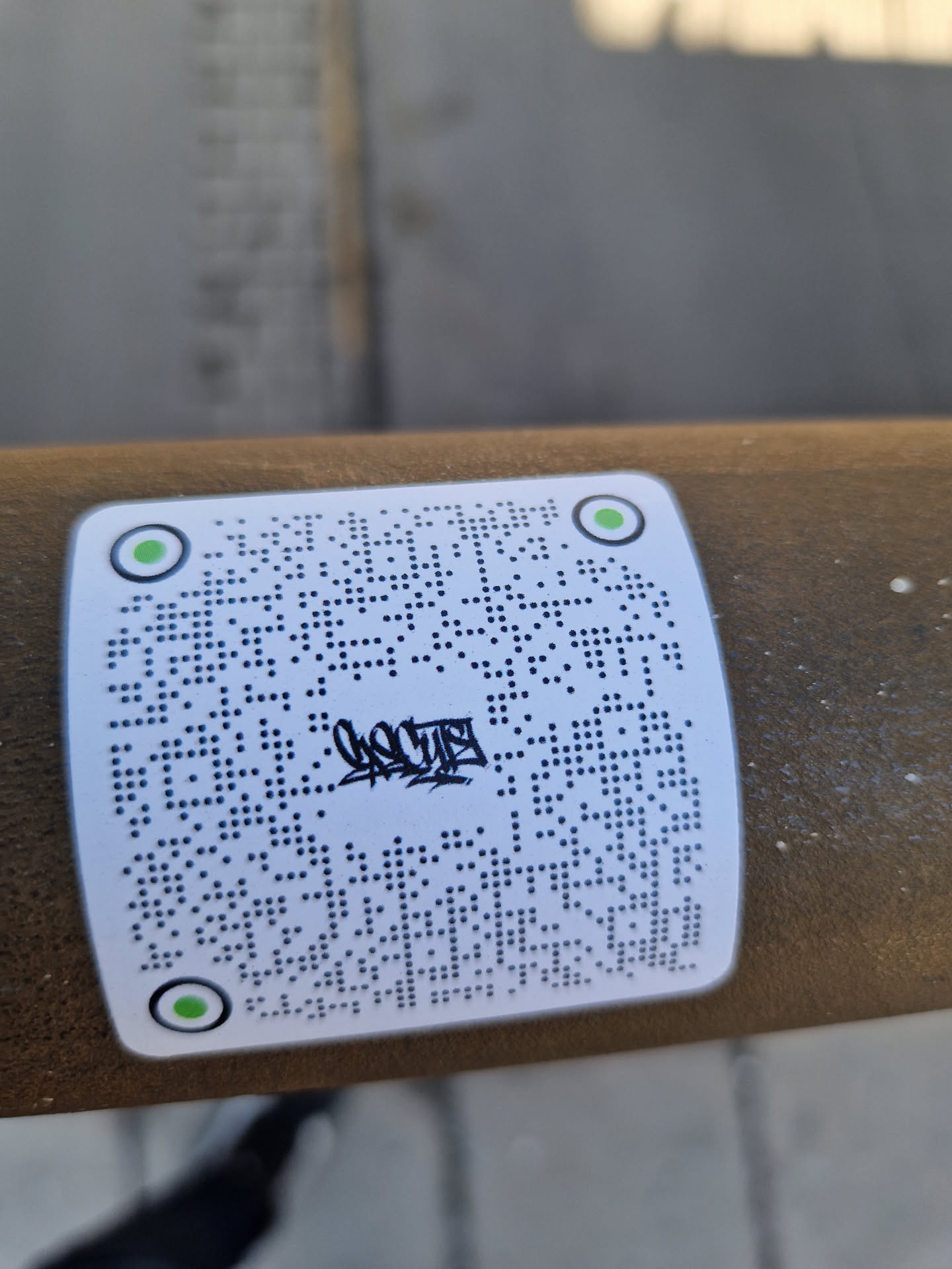 A white square sticker with a QR code and some graffiti-style text in the center. The sticker is affixed to a brown cylindrical object outdoors.  The QR code appears to be a standard QR code.