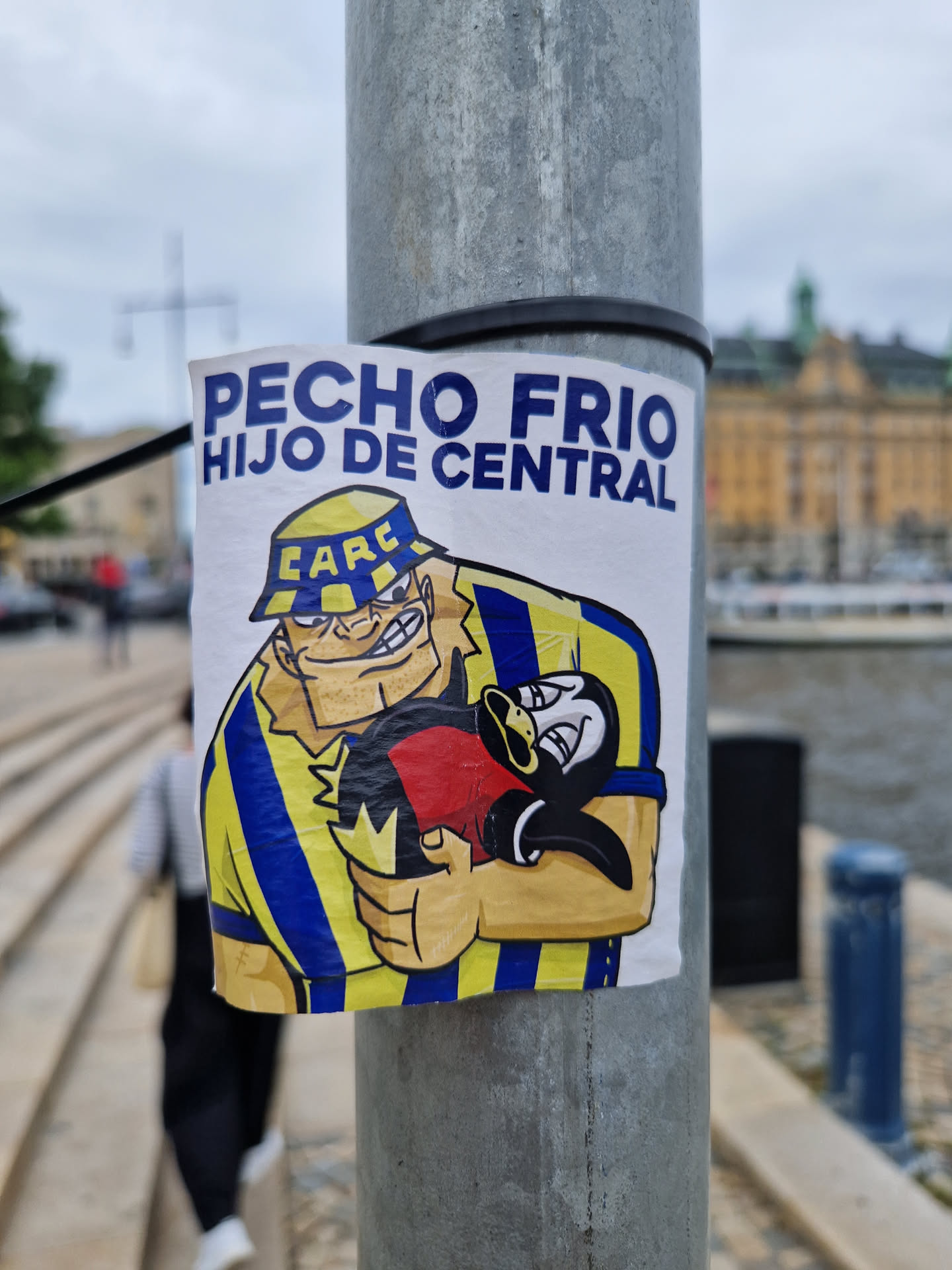 A sticker featuring a cartoon illustration of a strong man in a yellow and blue striped shirt holding a penguin. The text on the sticker says "PECHO FRIO HIJO DE CENTRAL".