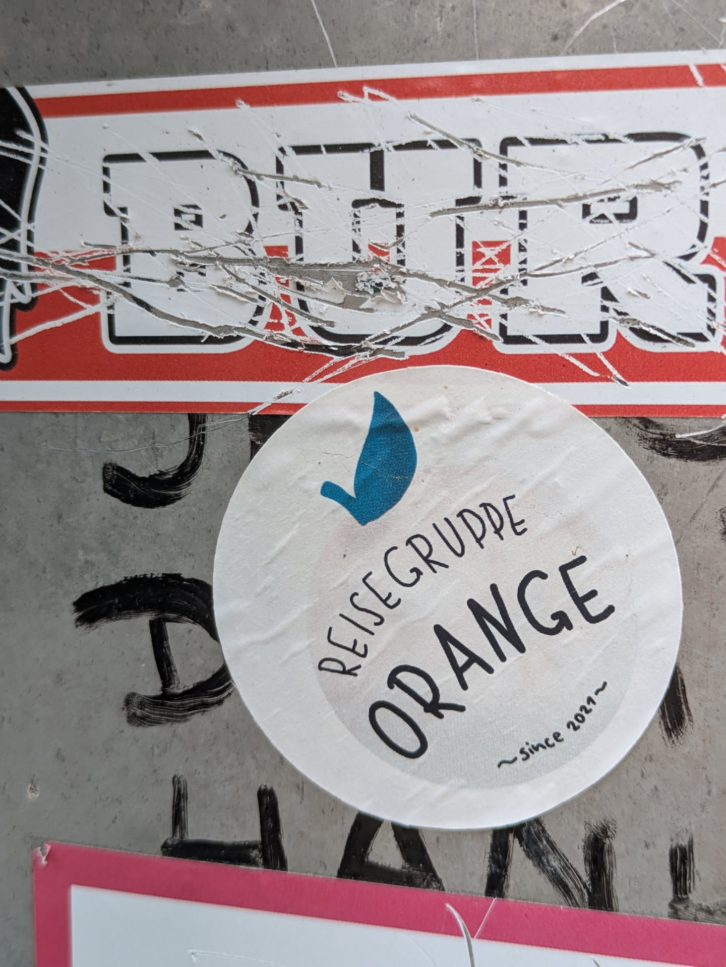 A circular white sticker with a prominent blue teardrop or stylized leaf symbol at the top. The German words "REISEGRUPPE" (travel group) are written in a curved line above the larger word "ORANGE", both in a black, somewhat informal or handwritten font. Below "ORANGE", the smaller text "~since 2021~" is present. The sticker appears slightly textured or wrinkled, suggesting it might be made of fabric or has been on the surface for some time.