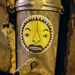 A round sticker featuring a stylized sun face with a serious expression. The sun is yellow with brown features, and the background is white. The sticker is attached to a rusty metal pipe. 