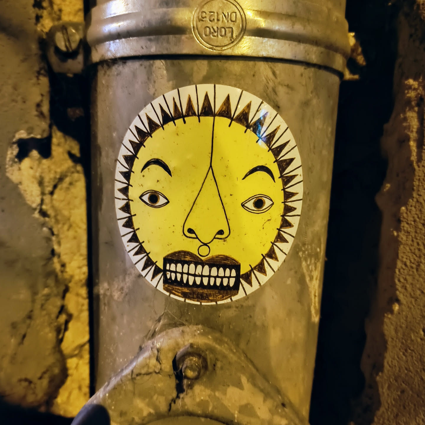 A round sticker featuring a stylized sun face with a serious expression. The sun is yellow with brown features, and the background is white. The sticker is attached to a rusty metal pipe. 