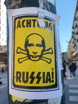 A sticker depicting Vladimir Putin's face superimposed on a skull and crossbones, with the words "ACHTUNG" and "RUSSIA!" in bold black letters on a yellow background. The sticker has a warning and protest tone against Russia.