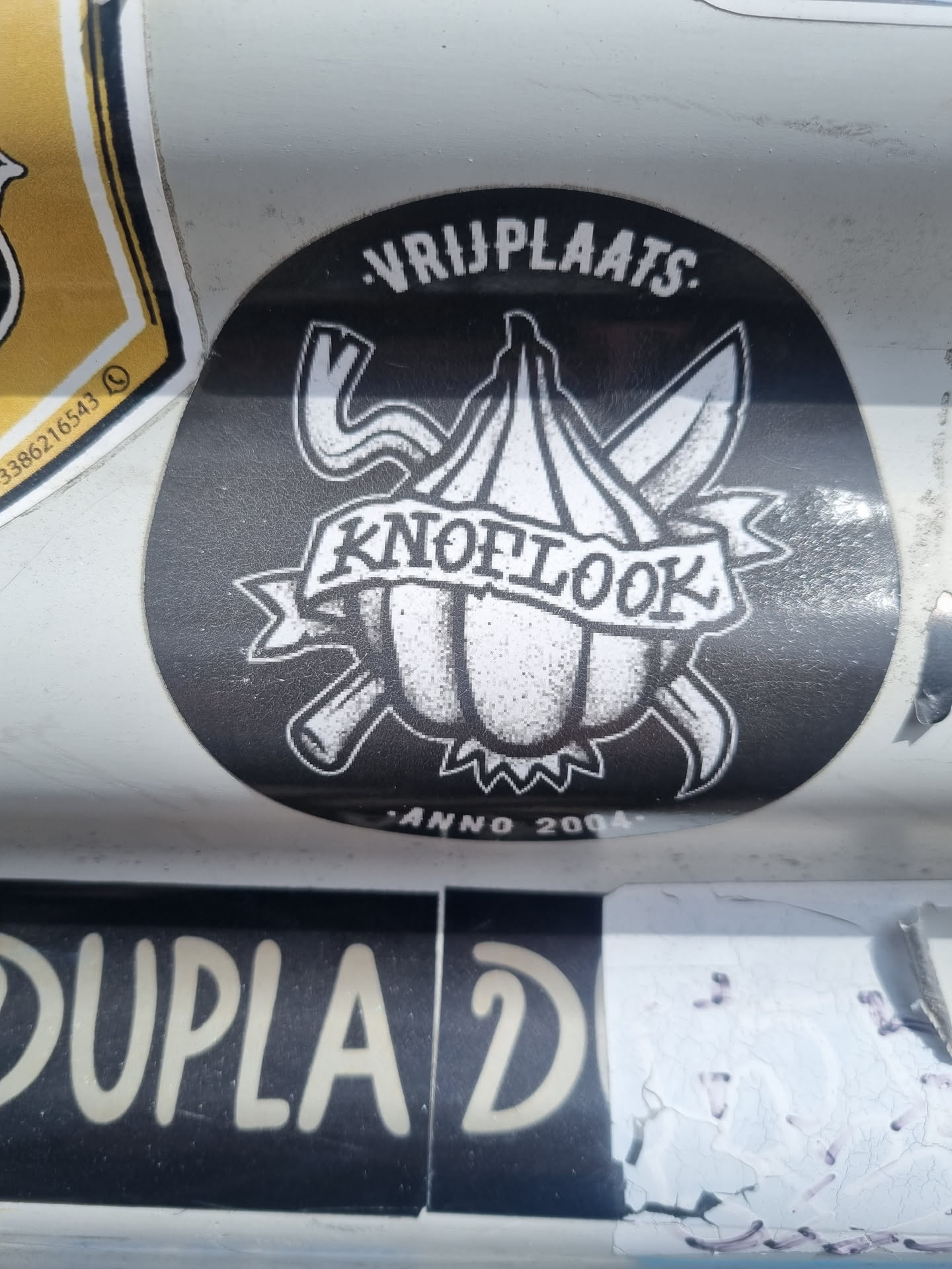 Black and white sticker with a drawing of a garlic bulb with two crossed knives behind it.  The text "VRIJPLAATS" is written at the top,  "KNOFLOOK" is on a banner under the garlic, and "ANNO 2004" is at the bottom.