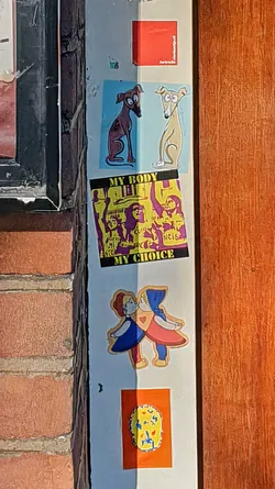 A series of stickers are affixed to a wall, showcasing a variety of designs. One features two greyhounds, one brown and one beige, against a light blue background. Below this is a "My Body My Choice" sticker with figures protesting in purple and yellow. Another sticker depicts two figures in blue and red kissing with a heart between them. The stickers are arranged vertically.